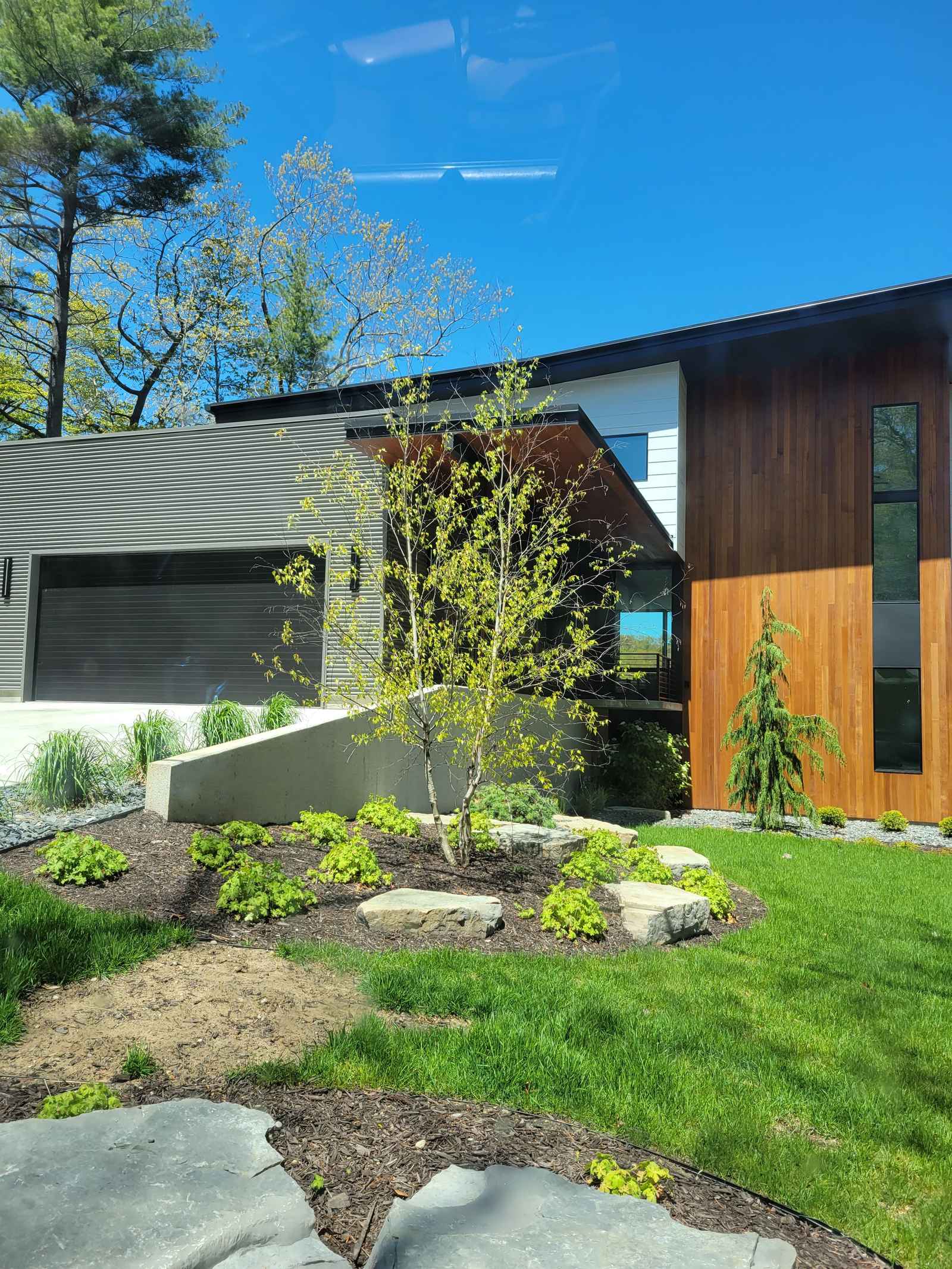 Completed modern custom home exterior with warm vertical wood siding, black metal accents, and a glass-paneled garage door
